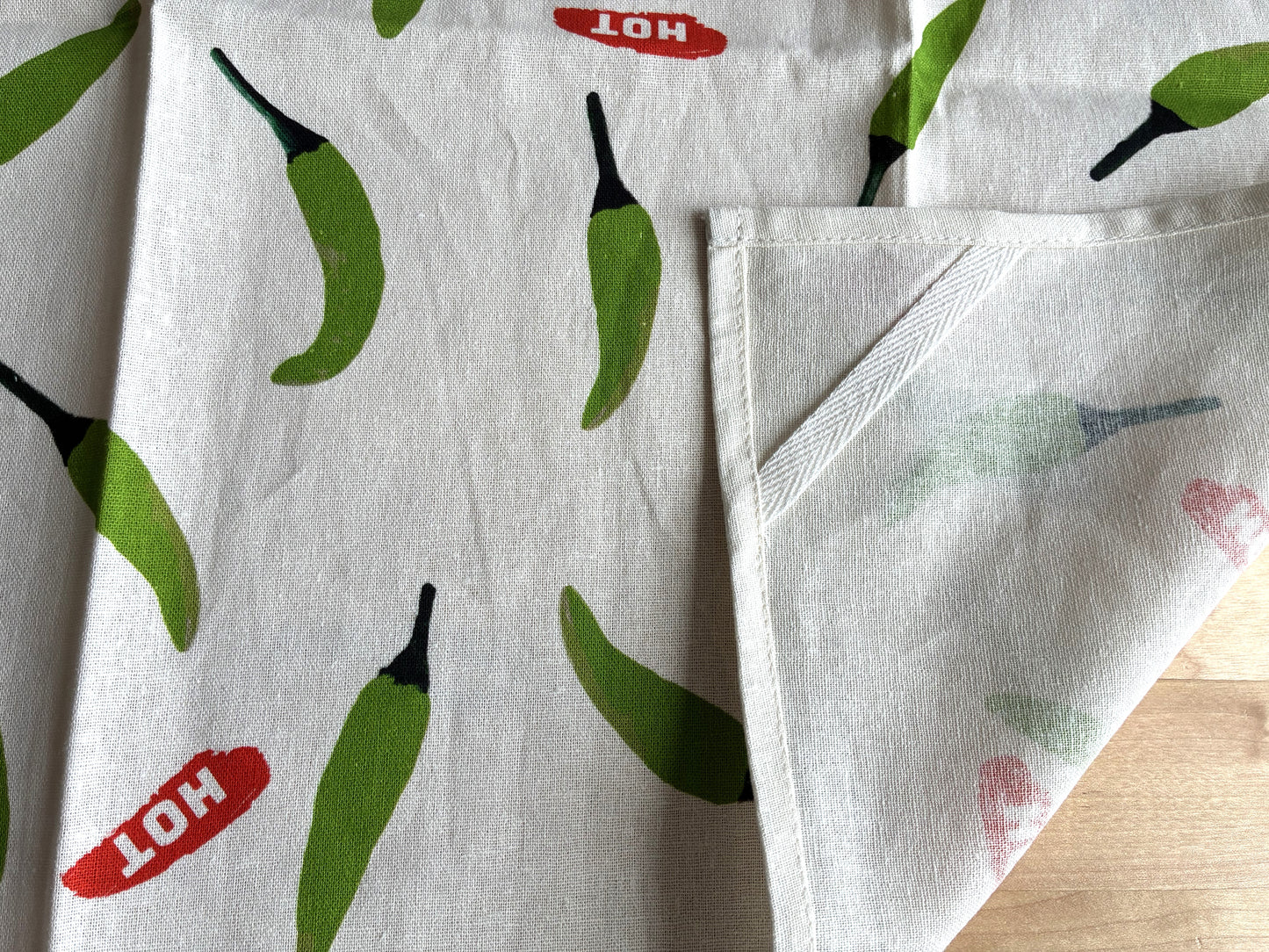 A close-up of a white fabric item, likely a tea towel or napkin, featuring a green chili pepper print and red 'HOT' labels, with one corner folded over to show the reverse side.