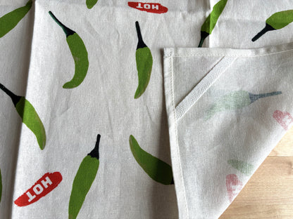 A close-up of a white fabric item, likely a tea towel or napkin, featuring a green chili pepper print and red 'HOT' labels, with one corner folded over to show the reverse side.
