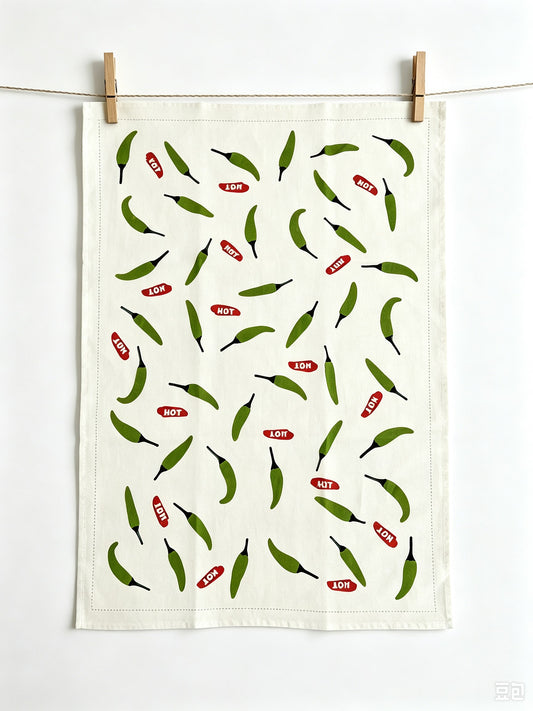 An off-white kitchen tea towel with a repeating pattern of green chili peppers and red 'HOT' labels, hanging from a string with two wooden clothespins against a white wall.